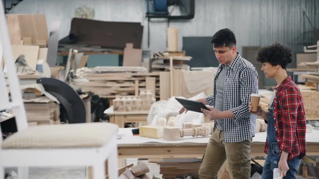 Business owners man and woman are talking walking in wood workshop holding take out coffee, papers and tablet. Start-up and communication concept.