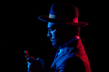 Young handsome model posing in a studio in a trendy neon light in a stylish suit and hat holding phone ,Internet threats concept.