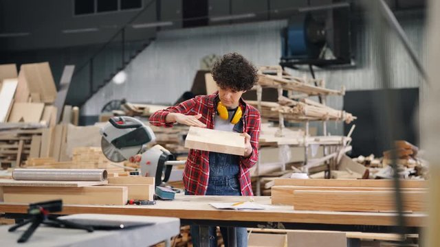 Careful female carpenter pretty girl is looking at piece of wood then polishing it with sandpaper working in workshop standing near worktable. People and job concept.