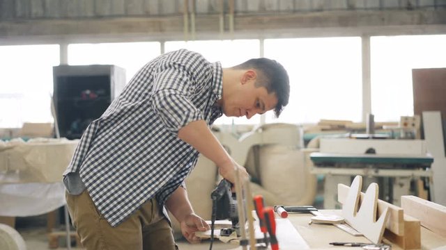 Handyman attractive guy is cutting wood with fretsaw using electric instrument in workshop then looking at woodware blowing dust. People and entrepreneurship concept.
