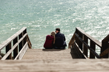 Couple in love sitting on stairs near ocean. Attractive pair enjoying ocean view on wooden path....
