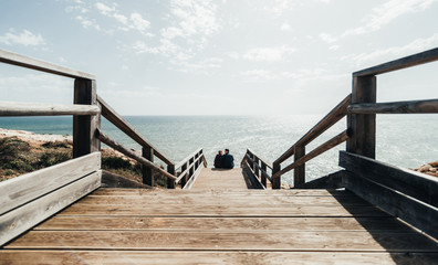 Couple in love sitting on stairs near ocean. Attractive pair enjoying ocean view on wooden path. Man and woman embracing and kissing on the beach in Portugal. Young family on a trip