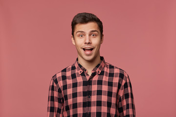 Portrait of young attractive happy amazed man in checkered shirt, looks at the camera with surprised expression, stands over pink background with wide open mouth.