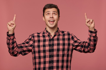 Fototapeta premium Young attractive happy amazed man wears in checkered shirt, looks up and wants to turn your attention to copy space over his head , isolated over pink background.