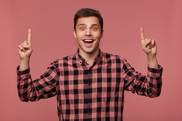 Naklejka premium Portrait of attractive happy amazed guy wears in checkered shirt, looks at the camera and wants to turn your attention to copy space over his head , isolated over pink background.