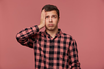 Portrait of young attractive man in checkered shirt, looks up, stands over pink background and touches head, looks tired and sad.