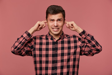 Obraz premium Portrait of young attractive guy in checkered shirt, looks at the camera, stands over pink background and covers ears from loud noise.
