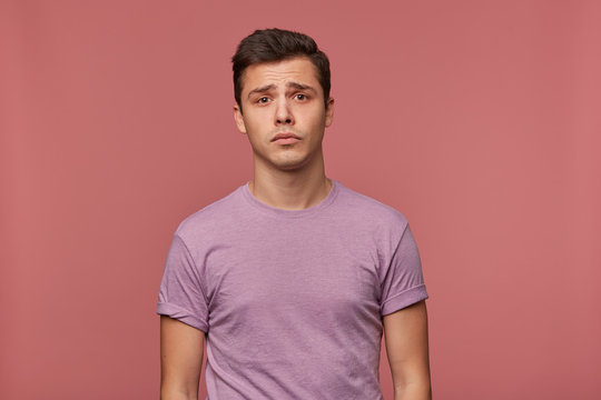 Portrait of young attractive boy in blank t-shirt, stands over pink background and looks sad and unhappy.
