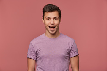 Obraz premium Portrait of cheerful handsome young man wears in blank t-shirt, looks at the camera with happy expression, stands over pink background.