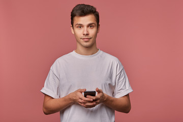Portrait of young handsome cheerful guy wears in blank t-shirt, looks at the camera with calm expression, stands over pink background and chatting with friend on the telephone.