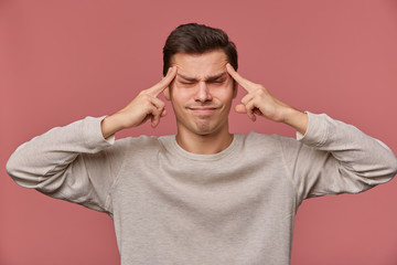 Photo of young attractive man in blank long sleeve, stands over pink background and shows headshot gesture with closed eyes, feels anhappy and migraineю