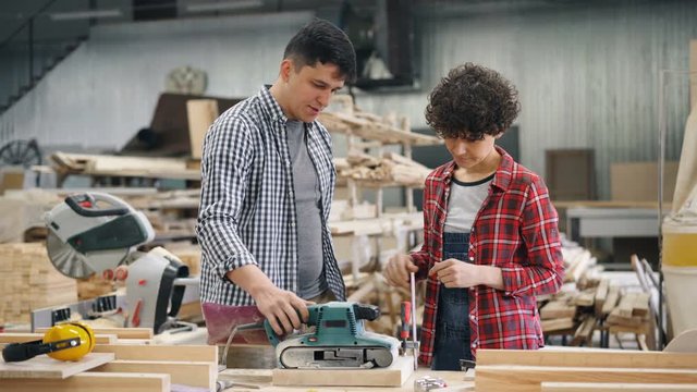Handsome guy professional carpenter is teaching new worker female apprentice to use polishing machine at factory. People, woodwork and communication concept.