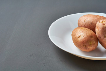 Three fresh raw potatoes on white ceramic plate on dark scratches concrete. Space for text