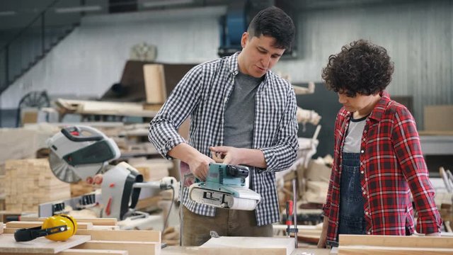 Guy professioal carpenter is teaching girl to use electric polishing machine in workshop talking holding instrument. People, factory and communication concept.