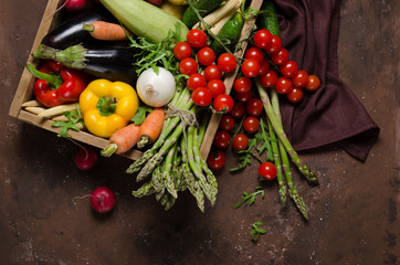 Box with farm vegetables on a dark background. Place for text. Cherry tomatoes, asparagus, broccoli, peppers, eggplants, zucchini, carrots and onions.