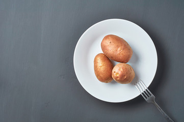 Three fresh raw potatoes on white ceramic plate with metal fork on dark scratches concrete. Space for text. Top view