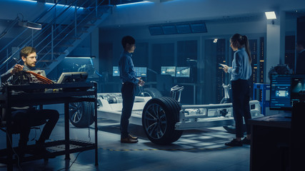Automotive Design Engineers Talking while Working on Electric Car Chassis Prototype. In Innovation Laboratory Facility Concept Vehicle Frame Includes Wheels, Suspension, Engine and Battery.