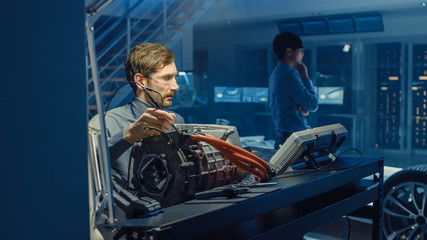 Professional Automotive Engineer in Glasses with a Computer and Inspection Tools is Testing an Used Electric Engine in a High Tech Laboratory with a Concept Car Chassis.