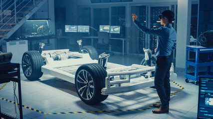 Automotive Engineer Working on Electric Car Chassis Platform, Using Augmented Reality Headset. In Innovation Laboratory Facility Concept Vehicle Frame Includes Wheels, Suspension, Engine and Battery.