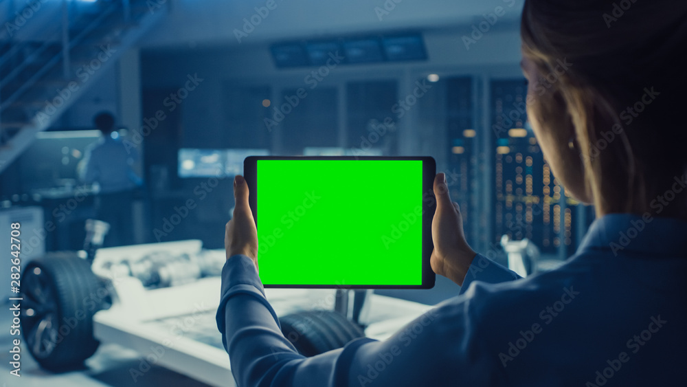 Poster female engineer holds a tablet computer with green screen mock up and looks at electric car chassis 