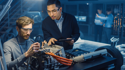 Two Professional Automotive Engineers with a Tablet Computer and Inspection Tools are Having a Conversation While Testing an Electric Engine in a High Tech Laboratory with a Concept Car Chassis.