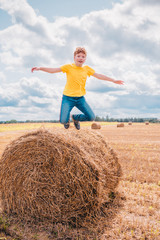 Naklejka na ściany i meble Red-haired Caucasian boy walks on a farm field among haystacks - wide angle and open aperture