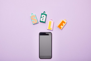 top view of colorful paper bags with sale lettering near smartphone with blank screen on violet
