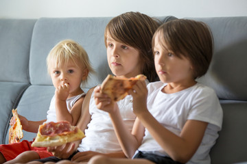 Cute children, sitting on couch, eating pizza and watching TV. Hungry child taking a bite from pizza on a pizza party