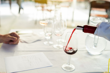 Man sommelier waiter pouring wine in the drinking glass at the testing ceremony event for the expert to test it and give marks about the quality to the degustation card