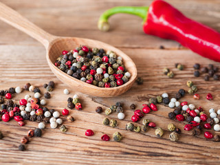Mix of peppercorns in wooden spoon on rustic background with red chili pepper. Red, black, green and white dry pepper in seeds.