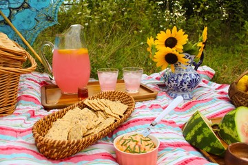 Beautiful country picnic in soft light meadow with basket sunflowers in pitcher watermelon pink lemonade pimiento cheese on colorful striped cloth, sun dappled 