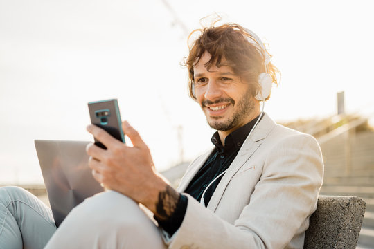 Portrait of smiling businessman with headphones and laptop looking at his mobile phone