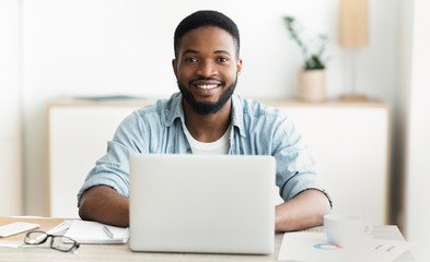 African guy using laptop, smiling and drinking coffee in office