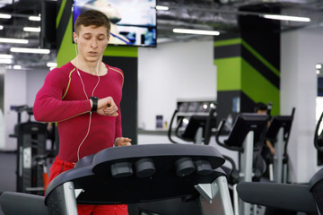 Full length portrait of muscular sportsman listening music in headphones and looking on the watches while using treadmill at modern gym