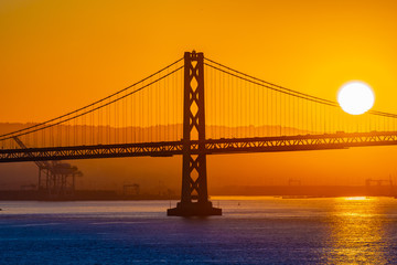 Beautiful summer morning sunrise behind mountain in California gives silhouette to the iconic Oakland Bay Bridge in San Francisco over the ocean.Famous travel location landmark in the west coast city.