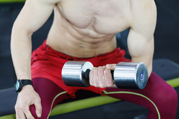 Close up view of strong topless man sitting and lifting dumbbells in the gym