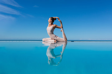 Charming woman yoga lover in overalls makes Mermaid Pose or Eka Pada Rajakapotasana asana by pool while relaxing at sea in sunny warm country. The concept of relaxation and better health. Copyspace