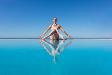 Beautiful young woman doing homukhasana sitting on the edge of the pool against the blue sky while relaxing on the beach. The concept of opening the chest and aligning the back. Copyspace