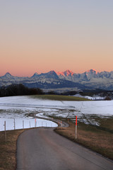 Pathway leading through winter hills on Gurten mountain with colorful evening sky and famous jungfrau mountain range (Eiger, Moench and Jungfrau).