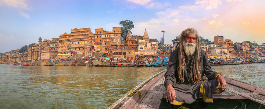 Indian sadhu on a wooden boat overlooking panoramic view of ancient Varanasi city architecture with Ganges river ghat