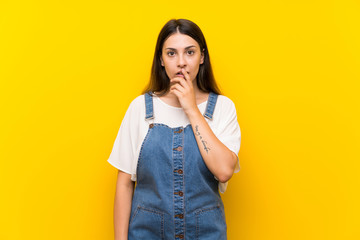 Young woman in dungarees over isolated yellow background surprised and shocked while looking right