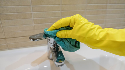 Woman in yellow rubber gloves cleaning a water tap. Housewife holding a rag. White sink. Modern bathroom. Cleaning company services. Housemaid washes the apartment.