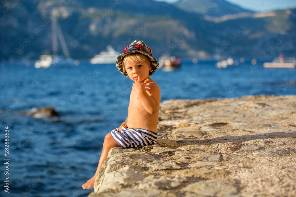 Sticker Sweet toddler boy with summer hat and sunglasses, sitting on the beach shore on sunset, enjoying