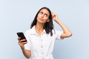 Young woman with a mobile phone over isolated blue wall having doubts and with confuse face expression
