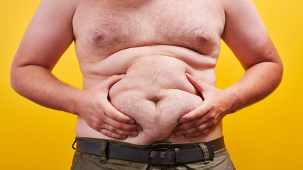 Man with naked body touches big fat belly on yellow background in studio closeup. Concept of...