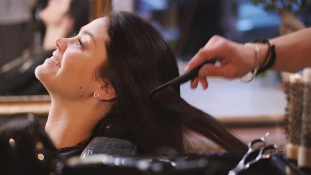 A woman in a hairdressing salon sitting near mirror and hairdresser brushing her hair with a comb. She is smiling 4k.