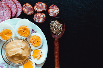 Slices of fresh appetizer foods placed on black background with copy space – Boiled egg halves, traditional salami and sausage bites with mustard bowl and wooden spoon with dried leaves