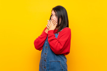 Young Mexican woman with overalls over yellow wall covering mouth and looking to the side