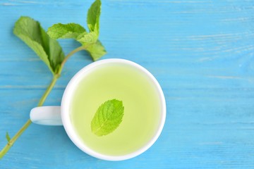 Curative mint tea in white porcelain cup with a green leaf of peppermint in tea and a sprig of fresh mint on blue textured wooden background. Healing hot mint tea. Calming peppermint beverage 