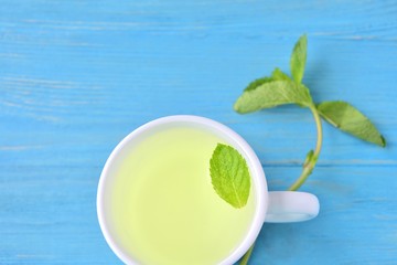 Curative mint tea in white porcelain cup with a green leaf of peppermint in tea and a sprig of fresh mint on blue textured wooden background. Healing hot mint tea. Calming peppermint beverage 